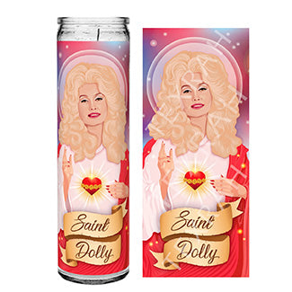 An artistic prayer candle design showing Dolly Parton styled as a saintly figure. She has big, voluminous blonde hair, a glowing halo behind her, and holds a heart with thorns. Her other hand is raised in a blessing pose. The text 'Saint Dolly' is written on a ribbon at the bottom, with a pink and red background.