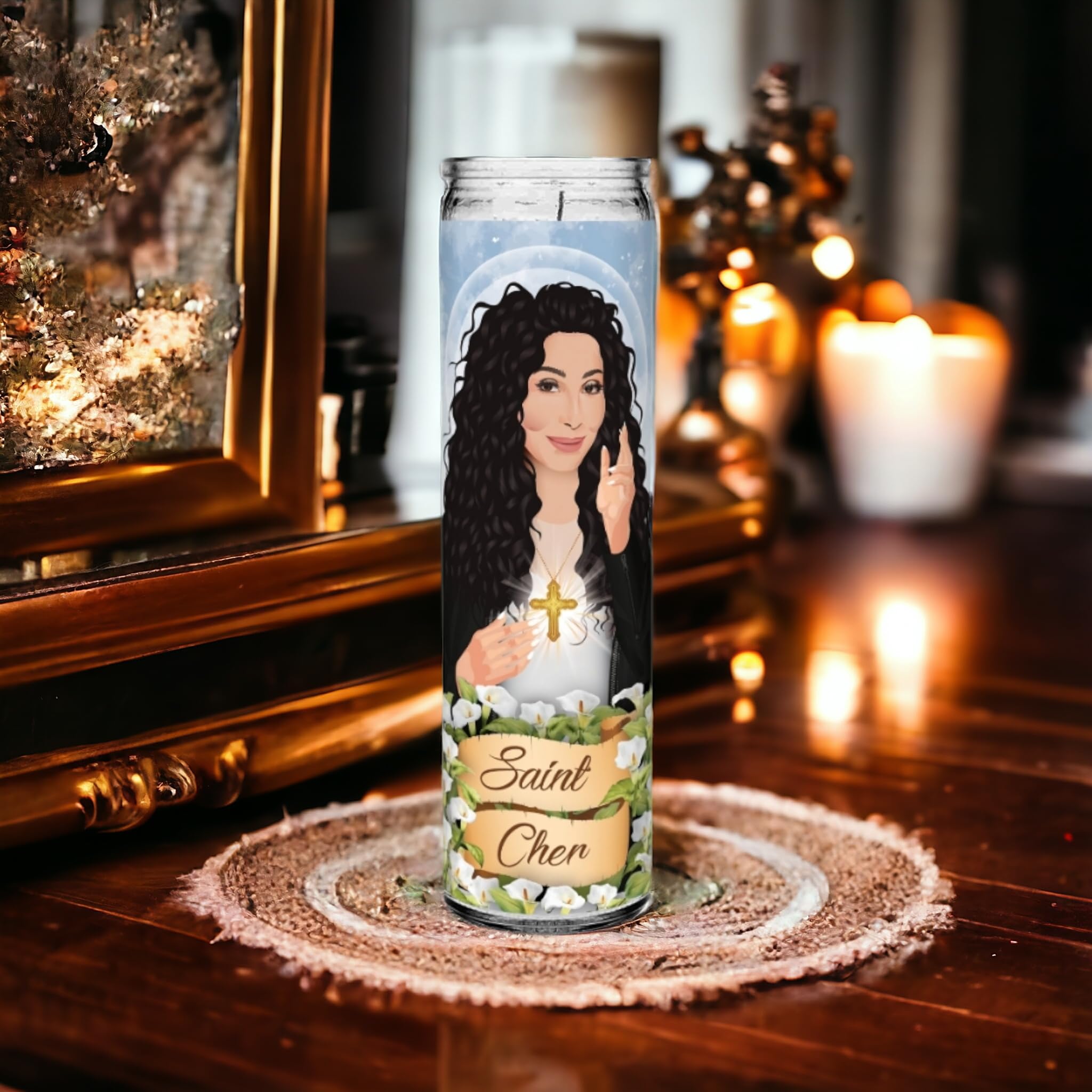 A photo of a 'Saint Cher' prayer candle sitting on a circular woven mat atop a wooden surface. The candle features an illustration of Cher with long, curly black hair, a glowing halo, and holding a cross over her chest. Her other hand is raised in a blessing gesture. The background includes a dimly lit room with soft, warm lighting and candles.