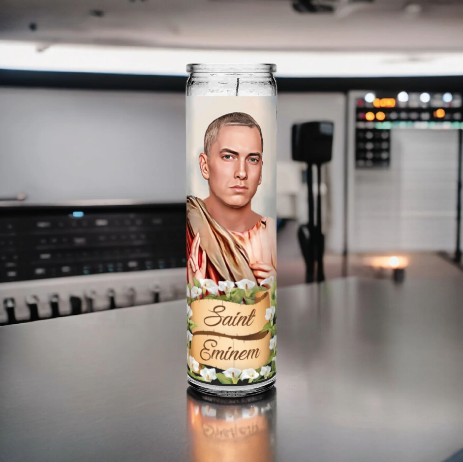 Saint Eminem prayer candle design featuring an illustration of Eminem as a saint, with short blonde hair and a serious expression. He is dressed in a robe, and the banner below reads 'Saint Eminem,' surrounded by white flowers. The candle sits on a desk in a recording studio.