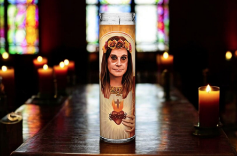 This image features the Saint Ozzy Osbourne Prayer Candle, set against a backdrop of glowing candles and vibrant stained-glass windows. The candle displays an artistic rendition of Ozzy Osbourne, wearing a floral crown, reminiscent of saintly depictions. The Sacred Heart symbol is prominently featured on his chest, blending Ozzy’s iconic rocker persona with a saintly reverence. The warm, ambient lighting and soft candle glow add a mystical and humorous touch to this tribute, perfect for honoring the Prince 